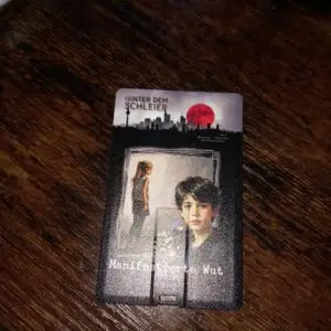 USB Stick mit dem Cover von Band 3 Der Sehr Flache Stick hat einen Ausklappbaren USB Stecker. Auf dem Cover ist im Hintergrund ein Mädchen das Vor Scherben steht im Vordergrund ein Junge der den Betrachter anschaut